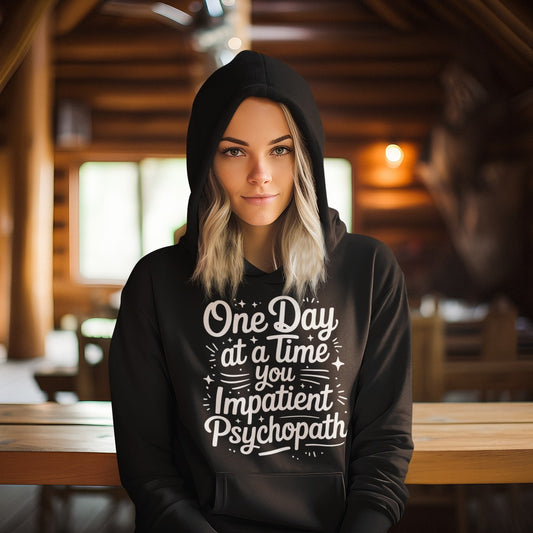 Woman wearing the black One Day at a Time You Impatient Psychopath hoodie, hood up, in a cabin.