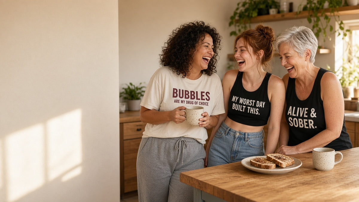 Three women laughing in a kitchen wearing SXD tees and tanks